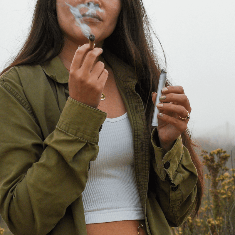 Person enjoying a smoke outdoors, holding a joint and a refillable lighter in a misty landscape.